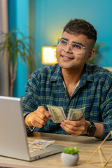 Happy Indian business man counting money cash and using laptop in home office. Successful freelancer guy getting bundle of money in office calculates dividends cash earnings profits finances. Vertical
