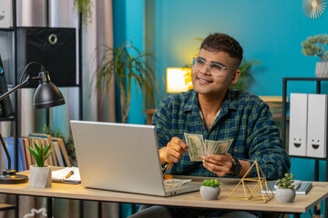 Happy Indian business man counting money cash and uses laptop in home office. Successful freelancer guy getting bundle of money in office calculates dividends cash earnings profits finances. Lifestyle