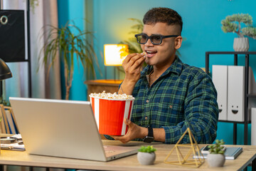 Business Indian man freelancer taking break from work wearing 3D glasses eating popcorn watching...