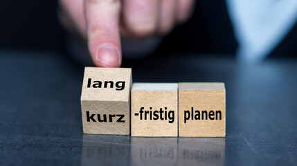Hand turns cube and changes the German expression 'kurzfristig planen' (short term planning) to 'langfristig planen' (long term planning).