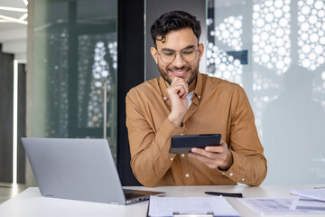 Financier auditor using calculator while working at office desk with laptop