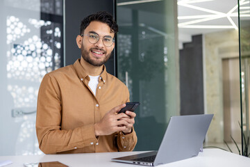Smiling young professional using smartphone and laptop in modern office environment