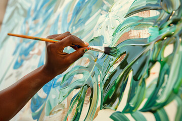 African female hand with paintbrush painting a picture of green monstera leaves in artwork studio.
