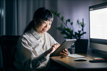 A happy japanese businesswoman at home office scrolling on tablet late at night.