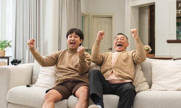 Cheerful Asian old man and middle aged son enjoy watching tv, celebrating good news. Smiling two male generations family feeling excited together, raising hands, resting on sofa in living room at home