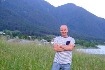 mature man, senior 60 years old standing in mountains above city, smiles, Alps are behind him,...