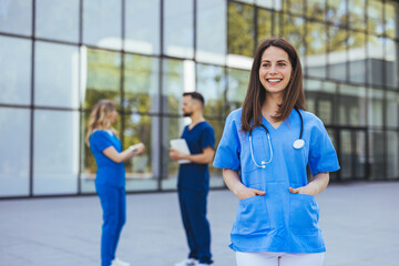 A smiling Caucasian female nurse in blue scrubs with a stethoscope stands confidently outside a...
