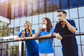 A trio of diverse healthcare workers in blue scrubs savor a relaxing coffee break outside a modern facility, rejuvenating with warm drinks and cheerful conversation.