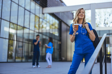 A cheerful woman in blue scrubs with a stethoscope takes a relaxed coffee break outside a medical facility, while colleagues converse in the background.