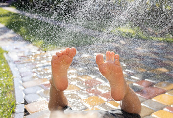 child lies barefoot on a paving slab under splashes of water from a hose or fountain. Child's feet in the sun and in the water. Summer fun. joyful mood. have fun. close up image
