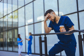 A weary male nurse in blue scrubs holds his forehead, expressing concern while holding coffee, with...