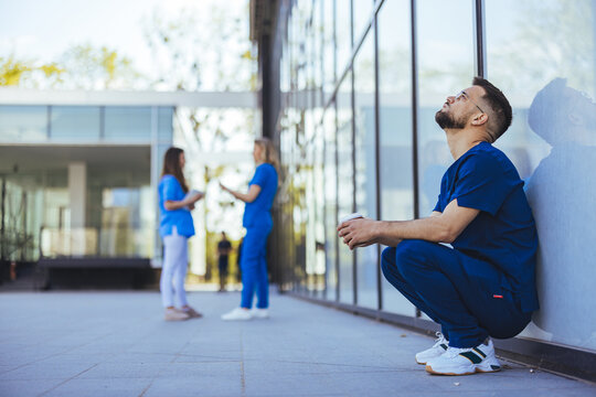 A weary male healthcare worker takes a moment to rest outside a hospital, his concerned colleagues engage in discussion behind him, reflecting the strenuous nature of their profession.