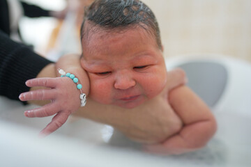 Mother bathing newborn baby in a modern kitchen, baby supported in a white tub, mother smiling...