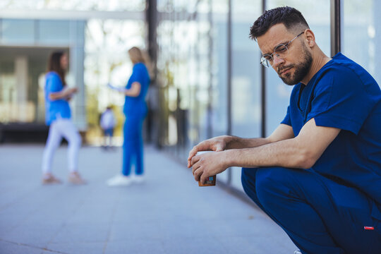 A Male Nurse Appears Contemplative While Seated On A Curb, His Scrubs Indicating Healthcare Work. In The Background, Fellow Nurses Engage In Discussion Near A Glass-walled Facility.