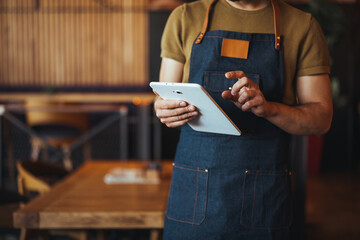 A cheerful male waiter in a denim apron uses a digital tablet to manage orders inside a cozy bar...