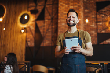 A joyful male waiter in casual attire, apron-clad, stands with a digital tablet ready to take...