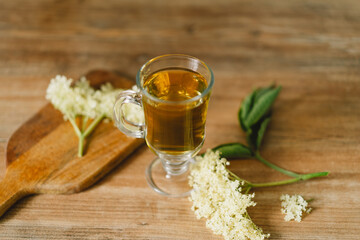 A clear glass mug filled with elderflower tea is placed on a rustic wooden table. Fresh...