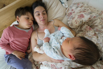 Mother holding her newborn baby in bed with her older child lying beside them, capturing a sweet...