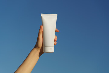 Female hand holding a white mockup tube of cream on a blue sky background. Concept of natural...