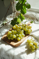 Fresh grapes on wooden charcuterie board on white table cloth in kitchen near morning window light with grape vine, Growing grapes indoors, Generative AI