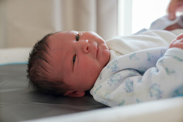 Newborn baby lying on a changing table, wearing a white onesie with blue and green patterns, gazing...