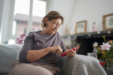 Senior woman in a grey shirt, sitting on a couch, holding a red smartphone, appearing thoughtful,...