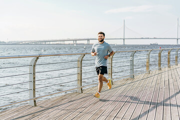 A man runs along a wooden boardwalk by the water, with a bridge and sailboats visible in the...