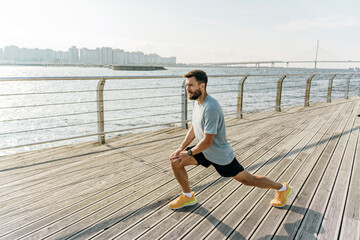 A man performs a lunge stretch on a wooden boardwalk by the water, with a bridge and city skyline...