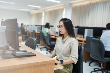 A woman sits at a desk in front of a computer. She is wearing glasses and is focused on her work. The room is filled with other people working on their computers, creating a busy