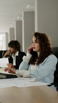 Caucasian pretty woman in sitting at the desc with a computer and working with documents while speaking on the phone in the office.