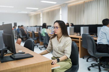 A woman sits at a desk in front of a computer. She is wearing glasses and is focused on her work....