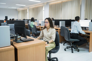 A woman sits at a desk in front of a computer. She is wearing glasses and is focused on her work. The room is filled with other people working on their computers, creating a busy