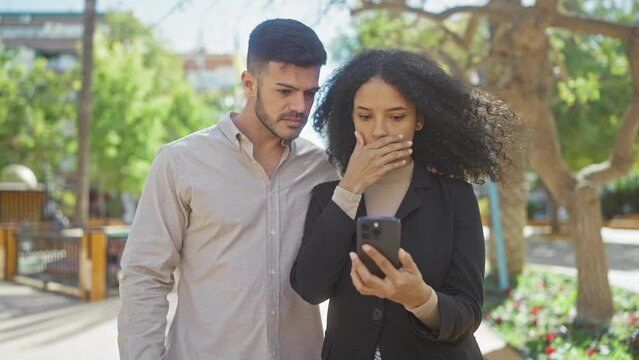 A man and woman react with shock and concern while looking at a smartphone together in a city park.