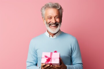 Portrait of a glad man in his 50s holding a gift while standing against pastel or soft colors...