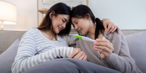 Couple smiling while looking at a positive pregnancy test on the couch at home. Women sharing a...