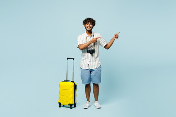 Full body traveler Indian man wear white casual clothes hold bag point aside isolated on plain blue...