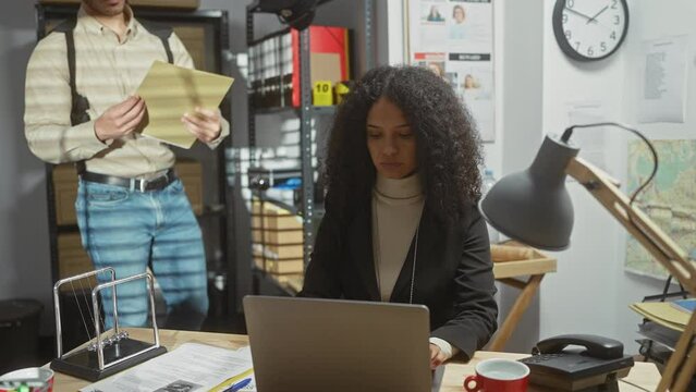 A woman detective analyzes evidence on a laptop while a man colleague shows her a document in a busy police station office.