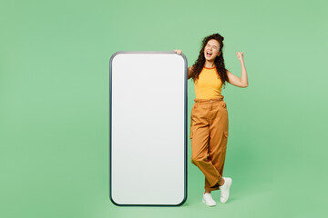 Full body young woman wear yellow tank shirt top big huge blank screen mobile cell phone smartphone with copy space mockup area do winner gesture isolated on plain green background. Lifestyle concept.