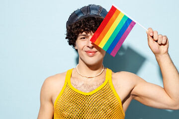 Young happy gay Latin man wear mesh tank top hat clothes hold cover eye with striped rainbow flag isolated on plain pastel blue cyan background studio portrait. Pride day June month love LGBT concept.