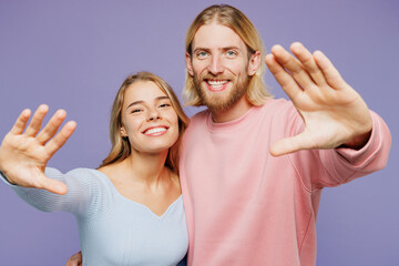 Young smiling happy cheerful fun couple two friends family man woman wearing pink blue casual clothes together stretch hands to camera isolated on pastel plain light purple background studio portrait.