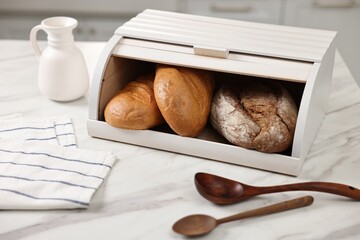 Wooden bread basket with freshly baked loaves and spoons on white marble table in kitchen