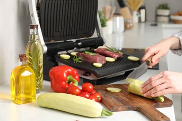 Woman cooking different products with electric grill at white wooden table in kitchen, closeup