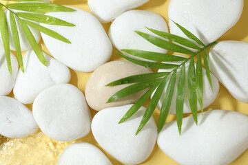 Spa stones and palm leaves in water on yellow background, flat lay