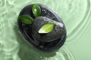 Spa stones and fresh leaves in water on light green background, top view