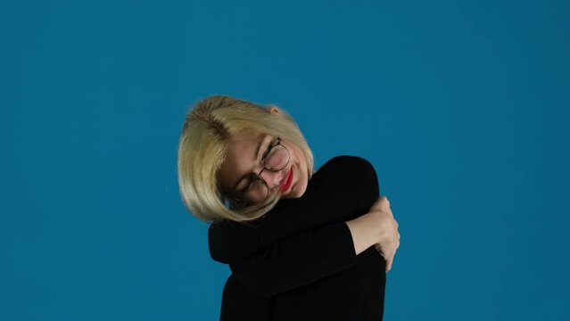 Businesswoman hugs own body expressing romantic feelings on blue background. Blonde office worker exudes happiness in tight embrace