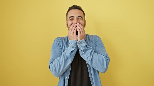 Young hispanic guy in denim shirt caught in gossip, laughing and covering mouth, posed against yellow isolated background, expressing embarrassed joy and shame