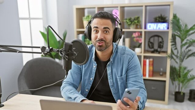 A handsome young hispanic man with a beard hosting a radio show indoors, using a microphone, laptop, and smartphone.