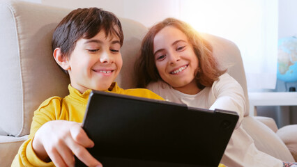 Two children are closely sitting on a sofa, engaged with a tablet
