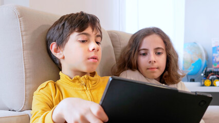 Two children are closely sitting on a sofa, engaged with a tablet