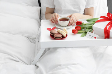 Tasty breakfast served in bed. Woman with tea, macarons, gift box, flowers and I Love You card at...